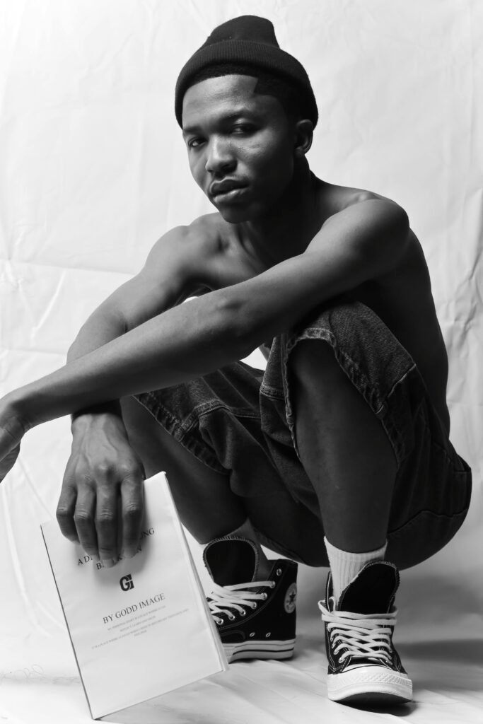 A stylish young man sitting in a studio wearing Converse shoes and holding a book, captured in black and white.